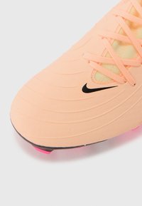 Chaussure de football Nike en pêche clair avec surface texturée, grand swoosh noir et accent rose sur la semelle. Dispose d'un design à lacets.