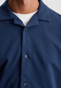 Close-up van een persoon die een gestructureerd marineblauw overhemd met knopen draagt over een wit geribbeld t-shirt met ronde hals.