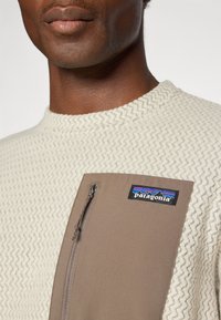 Gros plan sur une personne portant un pull texturé crème avec une poche zippée marron sur la poitrine, comportant un écusson du logo Patagonia.