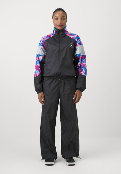 Femme debout portant des vêtements de sport noirs avec des motifs tie-dye roses et bleus sur les manches et une veste zippée avec des baskets.