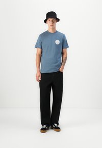 Blå bomull t-skjorte med et rundt hvitt logo, kombinert med svarte vide bukser og svarte joggesko med hvite detaljer.