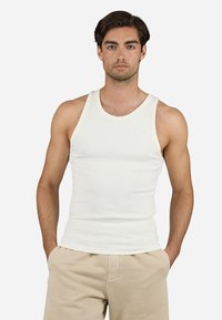 Hvit bomulls tanktopp med ribbet tekstur, med rund utringning og lysebeige detaljer. Liten logo på midten av fronten.