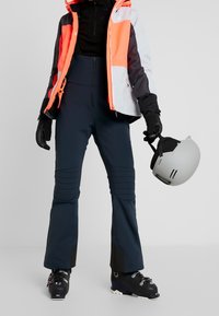 Skijacke in Orange, Grau und Schwarz mit einem Reißverschluss vorne; kombiniert mit marineblauen Skihosen mit strukturierten Seitenelementen und schwarzen Akzenten.