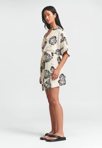 Květinové šaty v krémové barvě s modrými a oranžovými vzory, se zavinovacím designem, krátkými rukávy a látkovým páskem. Doplněné černými sandály.