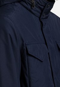 Mörkblå vadderad jacka med hög krage, tryckknappar och två framfickor, i ett slätt och lätt texturerat tyg.