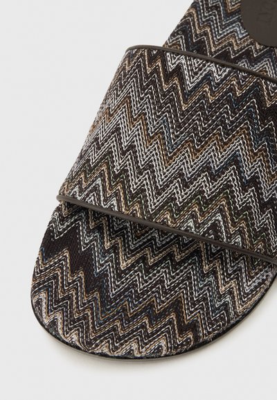 Chausson en tissu texturé avec un motif zigzag marron foncé, blanc et beige, bordé de couleur sombre, sur fond blanc.