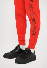 Sarkanas sporta bikses ar melniem "Emporio Armani" uzrakstiem sānos, kopā ar melnām ādas kurpēm ar biezām zolēm un melnām auklām.