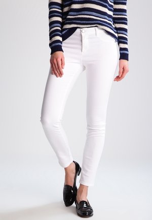 Vrouw draagt witte slim fit broek, marineblauwe gestreepte trui en zwarte lakleren loafers, staand met gekruiste benen.