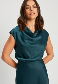 Zelena bluzica s kapuljačom i kratkimi rokavi, lahkoten satenski material, nabran pas, gladka tekstura in sproščen kroj.