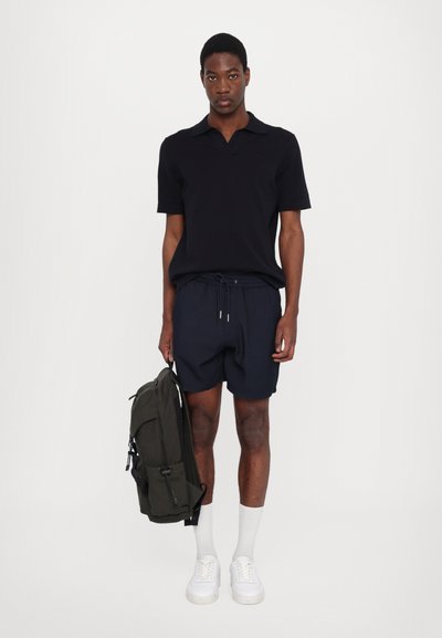 Polo azul-marinho e calções combinados com ténis brancos e meias de comprimento médio. O modelo segura uma mochila verde-escura. Design minimalista.