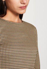 Haut à carreaux marron et noir avec un col rond, en tissu texturé et offrant une coupe suggérant un design structuré.