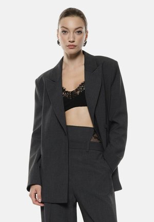 Blazer grigio su misura con una vestibilità rilassata, caratterizzato da ampi rever e due tasche frontali, indossato sopra un reggiseno in pizzo nero.