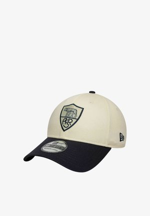 Casquette de baseball avec une calotte couleur crème et une visière noire, présentant un logo écusson noir brodé avec un loup et des lettres à l'avant.