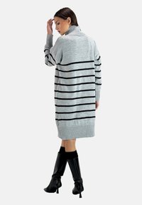 Robe pull en tricot gris avec des rayures horizontales noires, un col montant et des manches côtelées. Modèle portant des bottes à talons hauts noires.