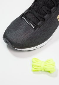 Chaussure de sport noire avec une tige texturée, une semelle blanche tachetée et des lacets noirs. Comprend un lot de lacets jaunes vifs.