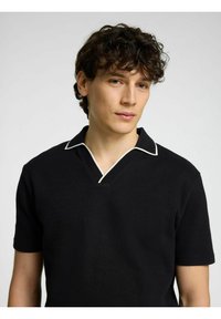 Črna polo majica s kratkimi rokavi, ki ima ohišje v obliki v-izreza in kontrastno belo obrobo ovratnika, izdelana iz mehkega, teksturiranega materiala.
