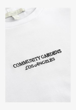 Bijela pamučna majica s crnim vezom na kojem piše: "ZAJEDNICE VRTOVI LOS ANGELESA." Okrugli dekolte i mekana tekstura.
