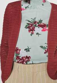 Rød strikket cardigan over en blomsterprint lysegrøn bluse med høj hals. Sammen med cremede stribede bukser.