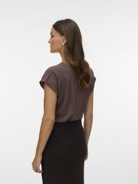 Γυναίκα με μακριά καστανά μαλλιά, που φοράει σκουλαρίκια με πέρλες, ένα καφέ μπλουζάκι με κοντά μανίκια και μια σκούρα φούστα με ρίγες, στραμμένη προς τα αριστερά με φόντο απλό.