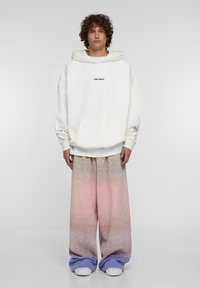 Felpa bianca oversize con tasca frontale e testo "LOST YOUTH" in nero. Abbinata a pantaloni larghi con gradiente dal rosa al blu chiaro e sneakers bianche.