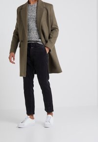 Manteau en laine vert olive, pull en tricot gris, pantalon en velours côtelé noir et baskets blanches. Les textures clés comprennent la laine et le velours côtelé.