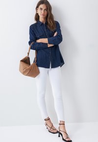 Camisa de botones de mezclilla oscura, jeans skinny blancos, bolso de cuero marrón claro y tacones negros de tiras. El fondo neutro enfatiza los detalles de la ropa.