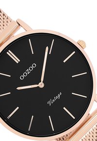 Cadran noir avec des index et des aiguilles en or rose. Le bracelet en maille complète le boîtier rond. Le texte indique "OOZOO Vintage."