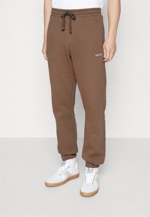 Brune joggingbukser med elastiske manchetter og snørelukning i taljen, båret med hvide sokker og hvide sneakers med gummisåler.