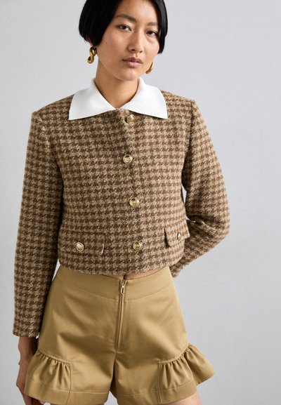 Chaqueta corta de patente marrón con botones dorados y cuello blanco, combinada con pantalones cortos color tan con un dobladillo con volantes y cierre de cremallera.
