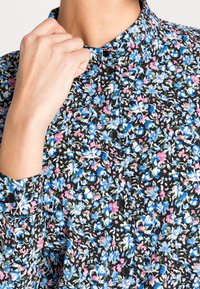 Main ajustant le col boutonné d'une chemise noire à motif floral dense en fleurs bleues, blanches et roses.