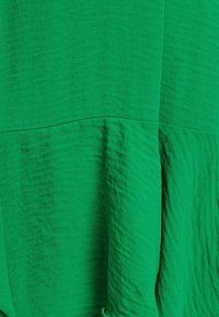 Zelená látka s texturovaným povrchem. Design má dvě části s viditelnými švy, což vytváří vrstvený vzhled.