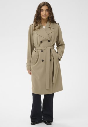 Beige trenchcoat med dobbeltradede knapper, bælte i taljen, sidelommer og vide ærmer, kombineret med mørke, viddejeans.