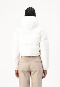 Vit kort vadderad jacka med hög krage och elastiska muddar, med en liten logotypdetalj. Bärs över beige högmidjade byxor.