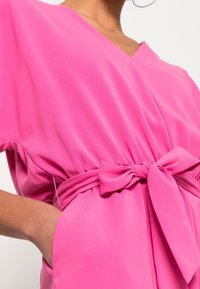 Vaaleanpunainen mekko, joka on valmistettu kevyestä kankaasta, V-pääntiellä, lyhyillä hihoilla, vyötäröllä nauhalla varustettuna ja sivutaskuilla. Sileä pinta.