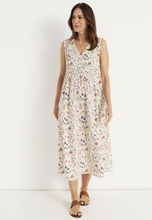 Woman wearing a sleeveless, ankle-length white dress with multicolored floral print and brown slip-on sandals, standing against a white wall.