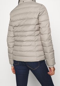 Světle šedá puff jacket s horizontálním prošíváním, vysokým límcem a knoflíkovými detaily. Materiál vypadá hladce a mírně leskle.