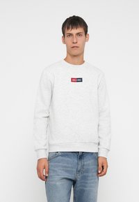 Grijze katoenen sweatshirt met ribbelmanchetten, ronde hals en een klein logo-embleem in rood en blauw, gedragen met vervaagde blauwe denim jeans.