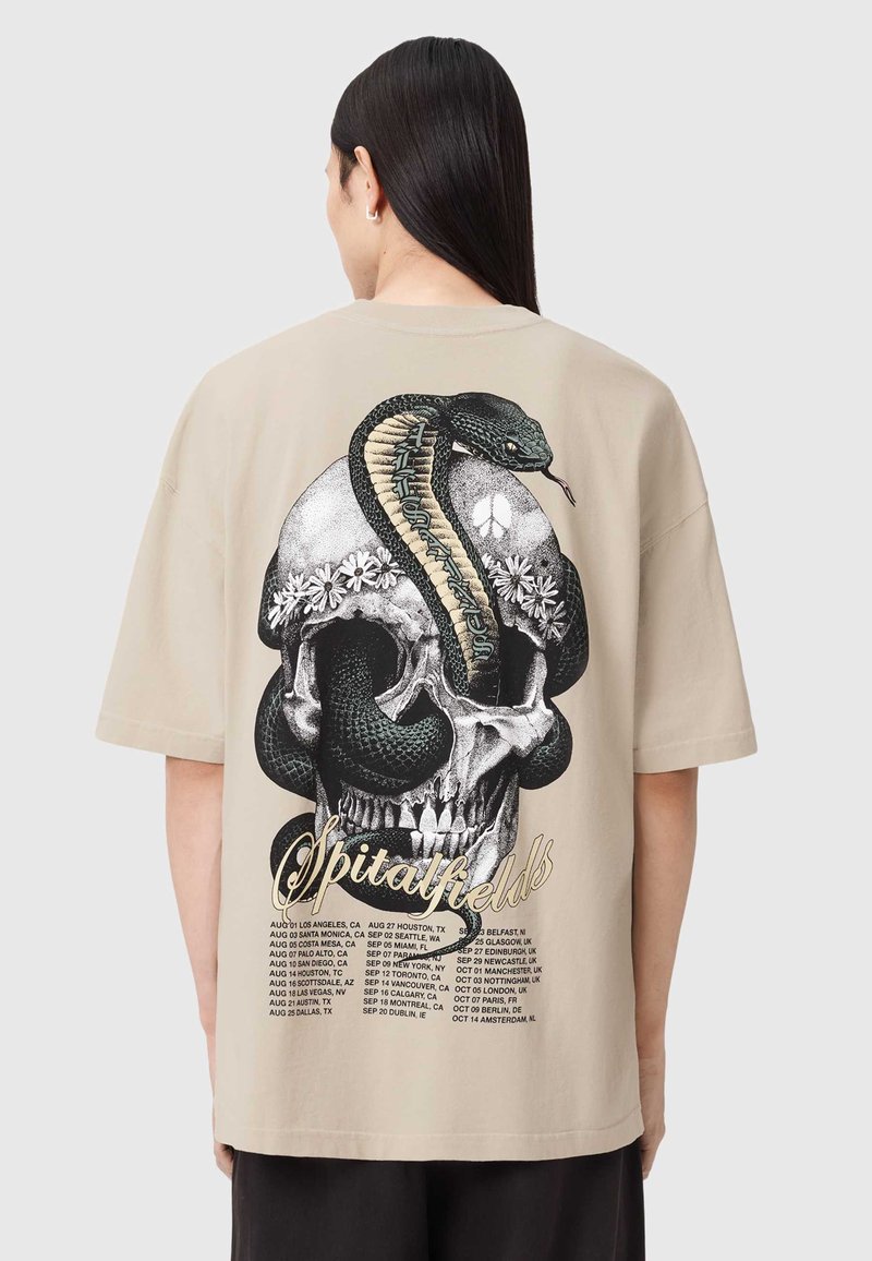 Camiseta oversize beige con un gráfico en negro de una calavera y una serpiente en la parte trasera, adornada con elementos florales y detalles del evento en la parte inferior.