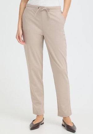 Personne portant un pantalon beige à jambe droite avec cordon à la taille, les mains dans les poches, un haut blanc et des ballerines noires fermées, debout à l'intérieur.