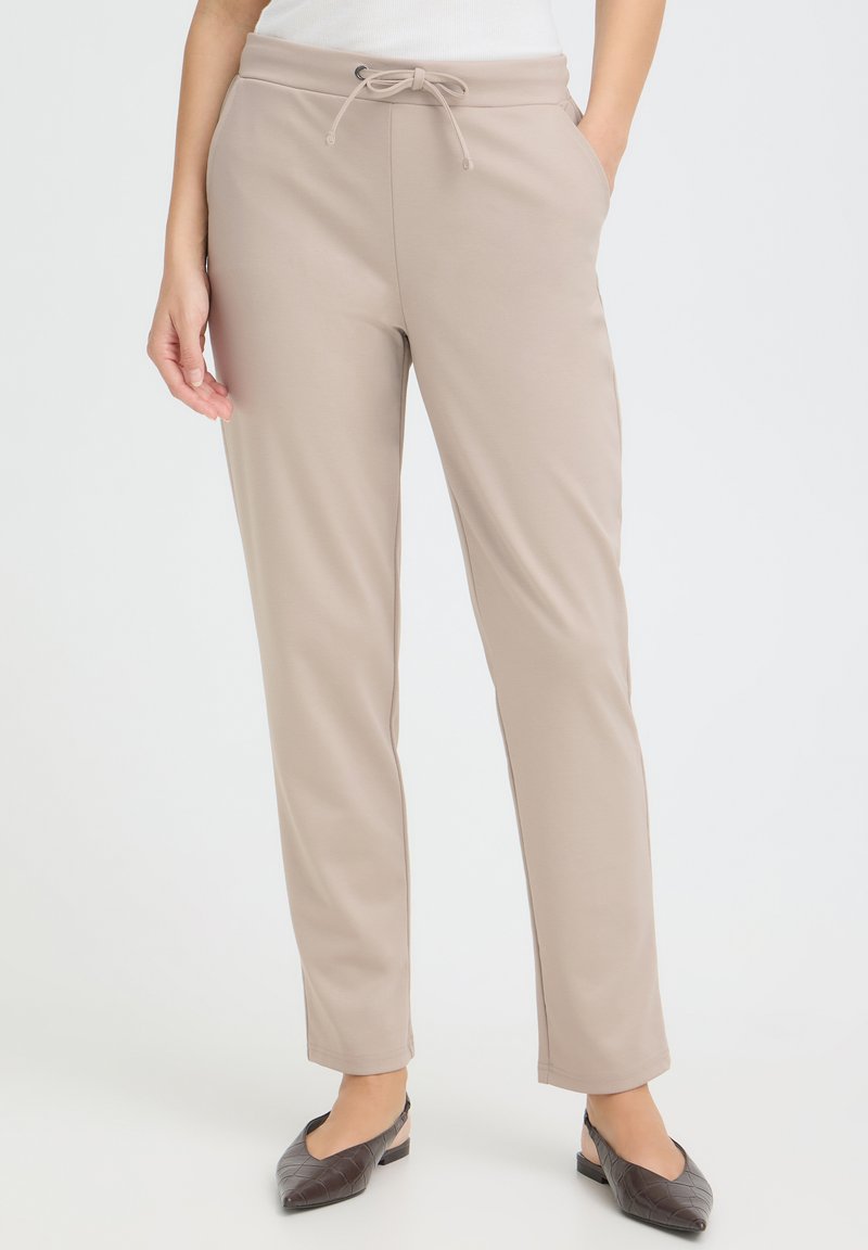 Personne portant un pantalon beige à jambe droite avec cordon à la taille, les mains dans les poches, un haut blanc et des ballerines noires fermées, debout à l'intérieur.