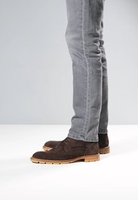 Bruine suède enkellaarsjes met een ronde neus, een lichtbruine rubberen zool en minimale veterdetails, gedragen bij lichtgrijze spijkerbroek.