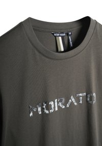 Camiseta verde oliva con cuello redondo y mangas cortas. En el frontal se presenta un gráfico camuflado de "MORATO" impreso en una fuente audaz.