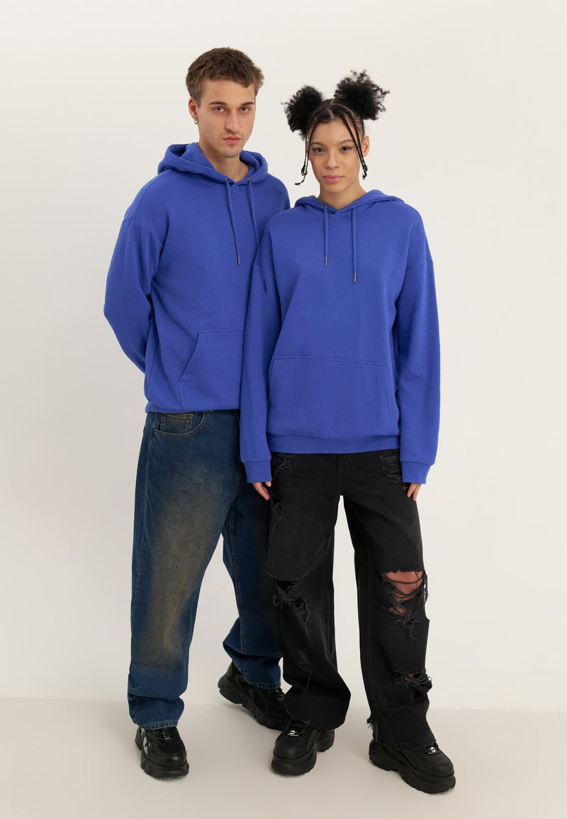 Blå hettegensere med frontlommer, brukt sammen med slitte svarte og blå jeans. Svarte støvler fullfører antrekket.