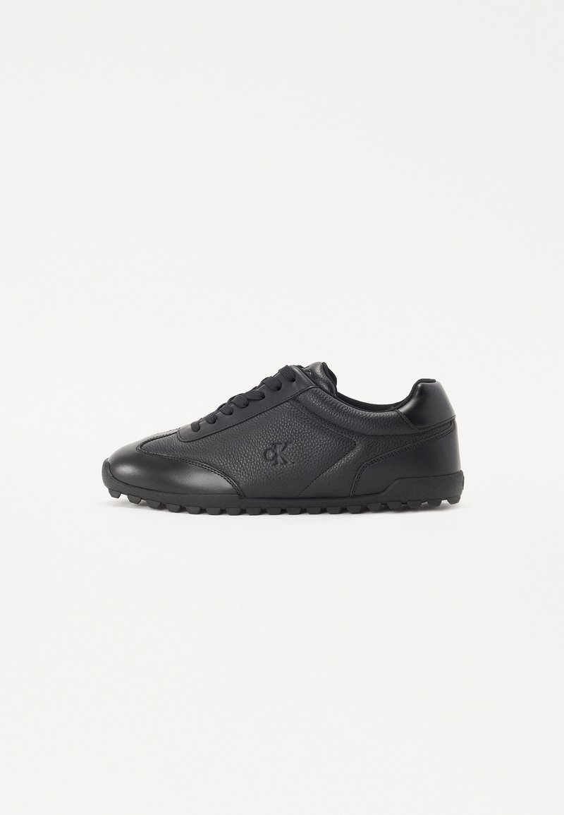 Black sneakers made of textured leather with smooth accents, featuring a round toe, flat rubber sole, and logo detail on the side.