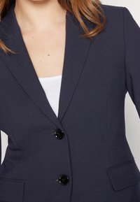 Granatowa marynarka z teksturowanej tkaniny, z wyraźnie zaznaczonymi klapami, dwoma czarnymi guzikami i przednimi kieszeniami, noszona na białym topie.