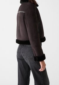 Brun shearling jakke med sort pelskant, kort design, lynlåslomme på siden og glatte læderdetaljer på ryggen og skuldrene.