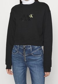 Person som bär en svart kortärmad sweatshirt med "cK"-logotyp och blå högmidjade jeans med sned dragkedja och "Calvin Klein Jeans"-märke.
