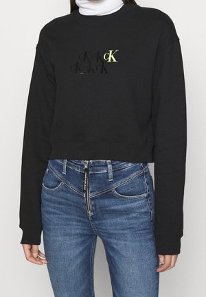 Persoon in een zwart cropped sweatshirt met "cK" logo en blauwe high-waisted jeans met diagonale rits en "Calvin Klein Jeans" label.