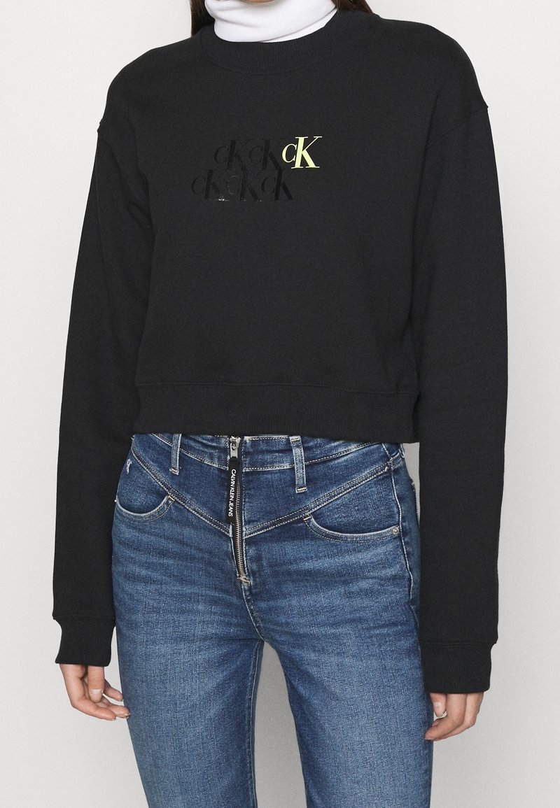 Person som bär en svart kortärmad sweatshirt med "cK"-logotyp och blå högmidjade jeans med sned dragkedja och "Calvin Klein Jeans"-märke.