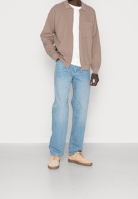 Uomo che indossa un cardigan beige con zip, una maglietta bianca, jeans azzurri chiari e sneaker beige in piedi contro uno sfondo bianco semplice.